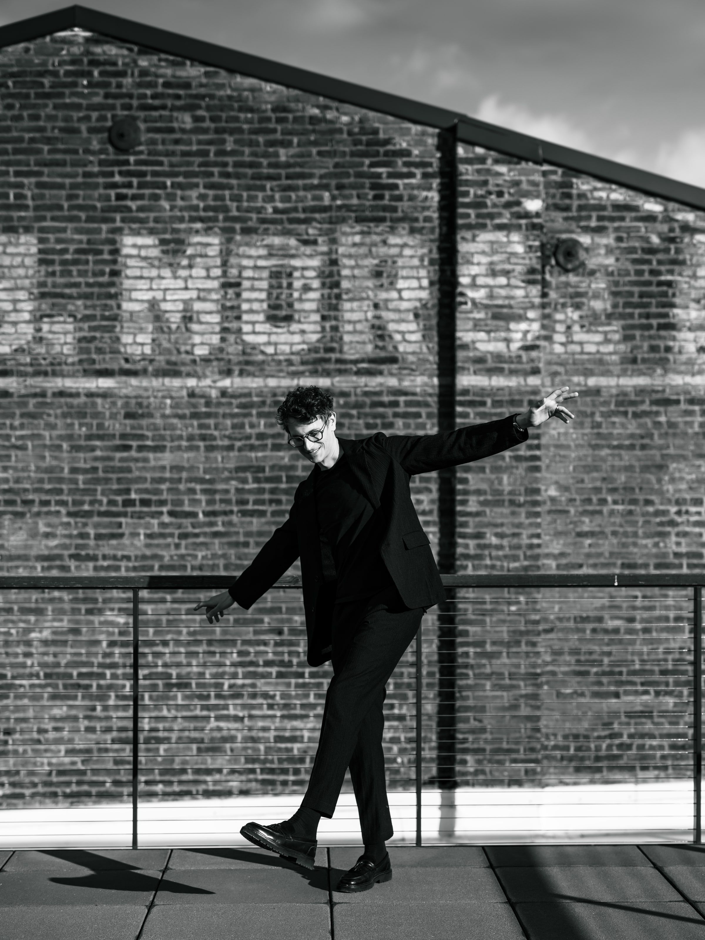 A black-and-white photograph of Matthew Claudel stepping across a rooftop with his arms out, in front of a brick wall with faded painted lettering.